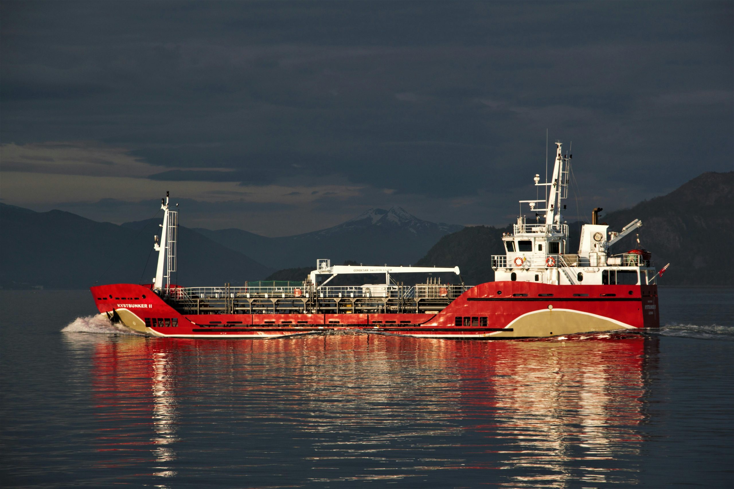 pexels-photo-7766101-7766101 A vibrant red cargo ship sails on the ocean at twilight with mountains in the background.
